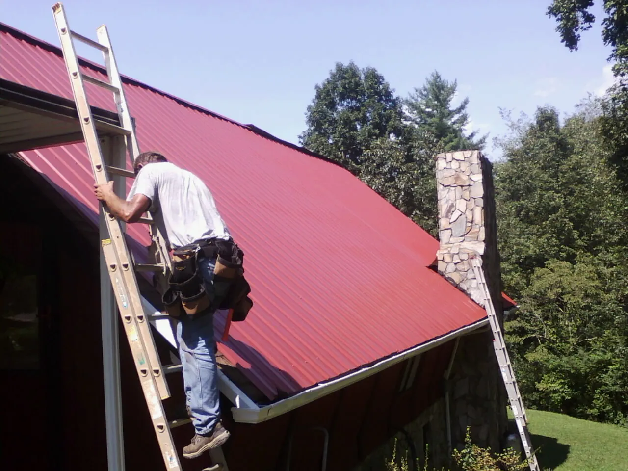 Roofer on a ladder beside a red metal roof and stone chimney — Soffit & Fascia Repair by Roofing Brothers in Berkeley Springs